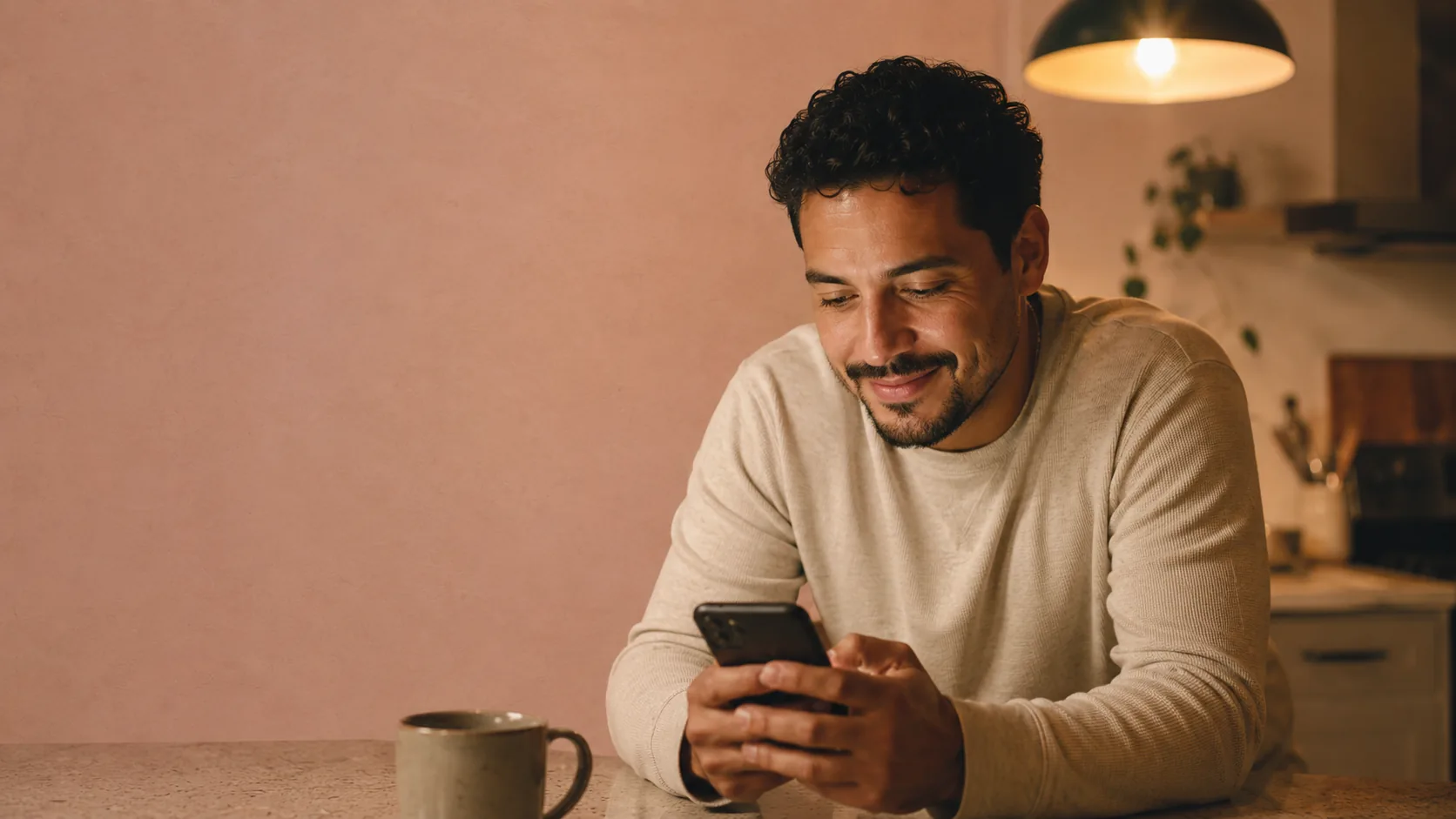 An adult son at his kitchen counter on a Saturday night, holding his phone with a hopeful smile after ordering a Mother's Day song
