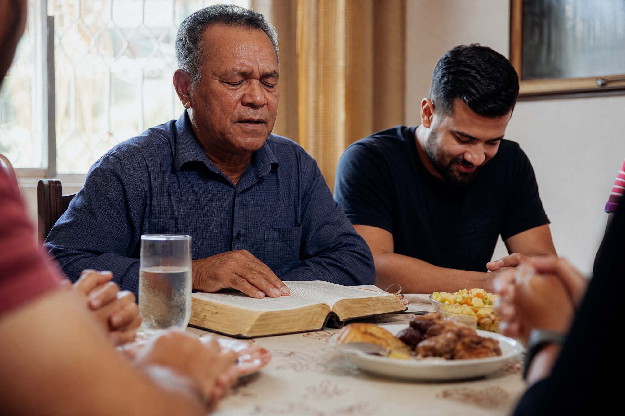 Pai evangélico brasileiro orando com a Bíblia aberta no almoço de domingo ao lado do filho, representando a homenagem musical de Dia dos Pais