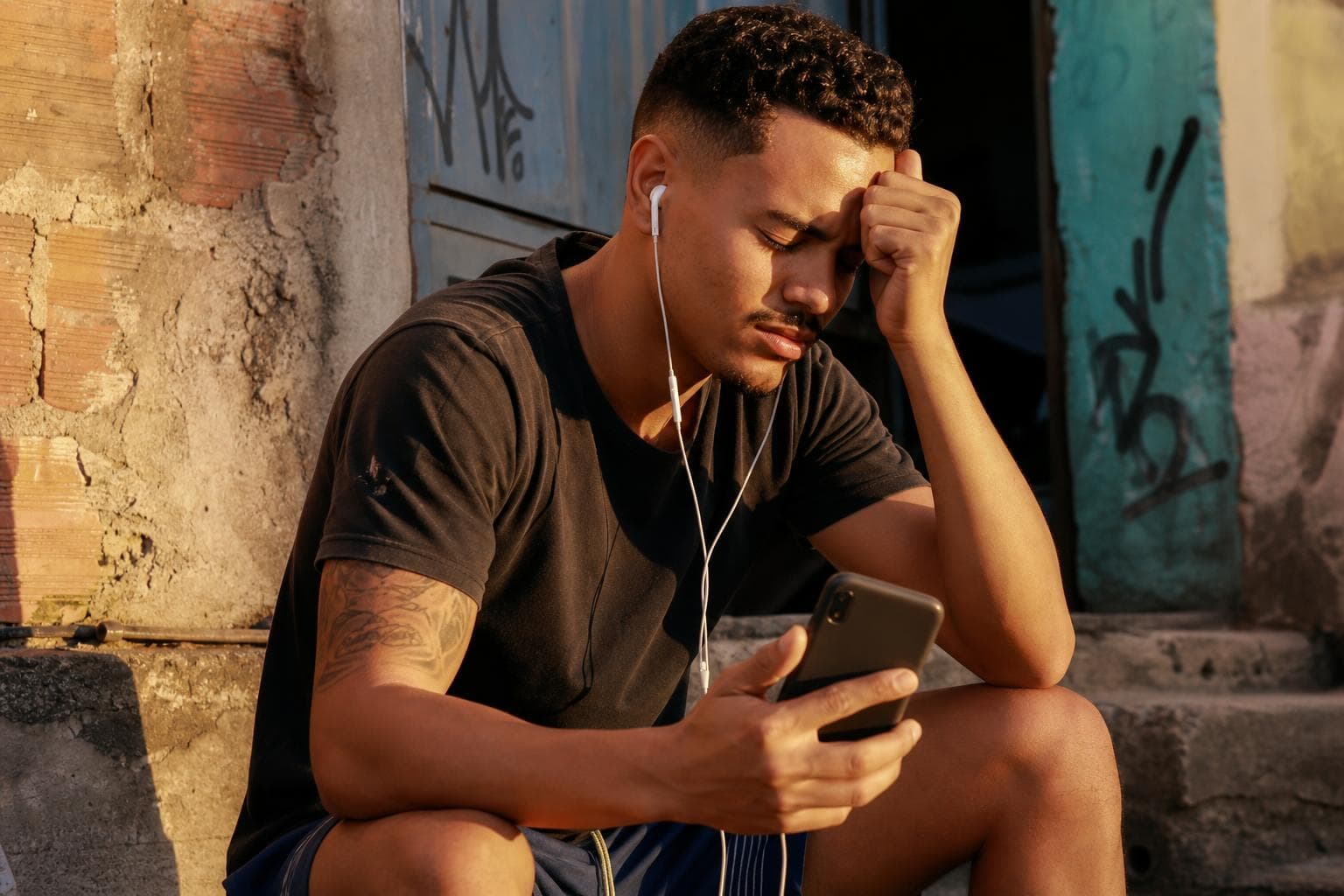 Homem jovem ouvindo música de homenagem para amigo falecido, sentado em degrau de casa na periferia