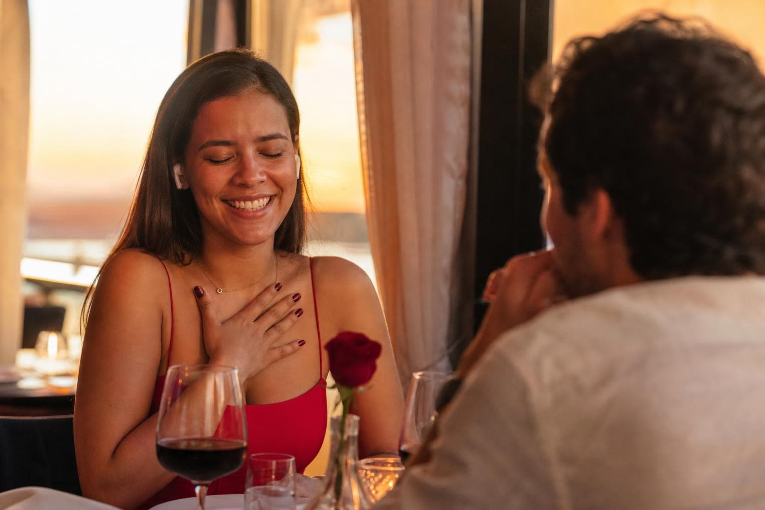 Casal jovem brasileiro em jantar romântico, ela ouvindo música personalizada com fones de ouvido, expressão de surpresa e emoção