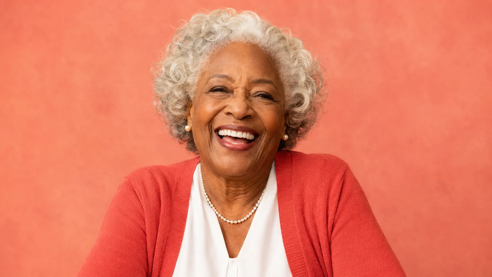 Portrait of Patricia — 75-year-old Black American grandmother with white-grey curls, big radiant laugh, deep coral cardigan